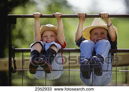 Stock Image - Agriculture - Young farm girl and boy wearing cowboy hats and rubber boots swing on a metal gate, hanging by their hands and legs and laughing / Northwest Missouri, USA.. Fotosearch