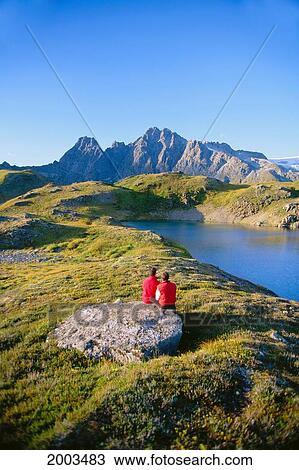 Stock Image - Alaska, Juneau, View Of Couple Sitting Beside Lake In Snow Capped Mountains, Blue Sky. Fotosearch