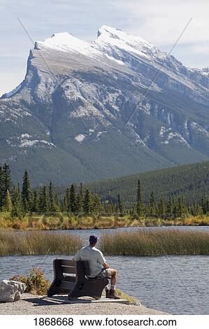 Stock Photo - Alberta, Canada; A Male Hiker Sits On A Bench Looking At Mount Rundle And Vermilion Lakes. Fotosearch