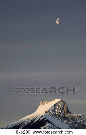 Alberta, Canada; Snow Covered Mountain Peak And Half Moon At Sunrise In Kananaskis Country View Large Photo Image Stock Photo - Alberta, Canada; Snow Covered Mountain Peak And Half Moon At Sunrise In Kananaskis Country. Fotosearch