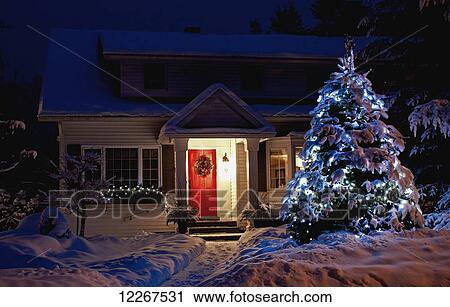 Stock Image - An illuminated tree in front of a house with a red door at Christmas time; Knowlton, Quebec, Canada. Fotosearch