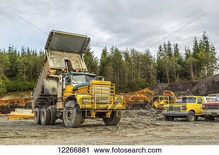 Stock Image - An old dump truck is positioned in a gravel yard with box raised up. Lumber and other equipment are shown in the background of the work yard against the forest; Port McNeill, British Columbia, Canada. Fotosearch