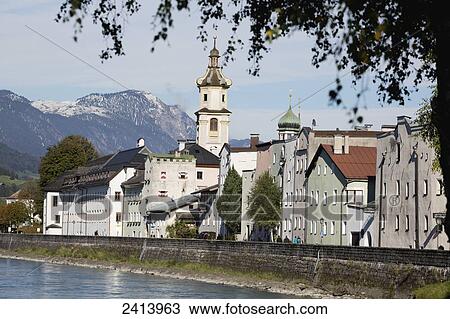 Stock Image - An old town along a river with stone barrier and church steeple with mountains and blue sky in the background; Rattenburg, Austria. Fotosearch