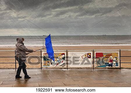 Stock Image - An Umbrella Blows Away As A Person Walks On A Promenade Along The Water On A Rainy Day; Redcar, Teesside, England. Fotosearch