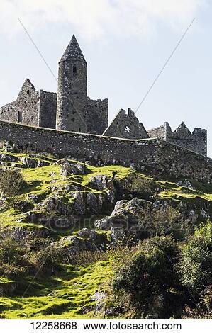 Stock Photo - Ancient stone ruin with stone wall, turret and church on rocky grassy hillside with blue sky and clouds; Cashel, County Tipperary, Ireland. Fotosearch