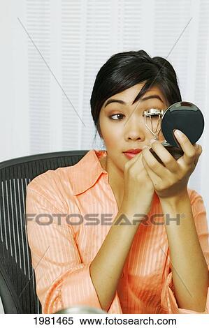 Stock Photography - Asian Businesswoman Sitting At Her Office Desk Using Eyelash Curler.. Fotosearch
