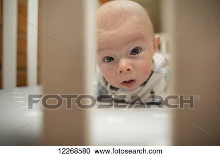 Stock Image - Baby holding its head up and looking through the bars of a crib, USA. Fotosearch