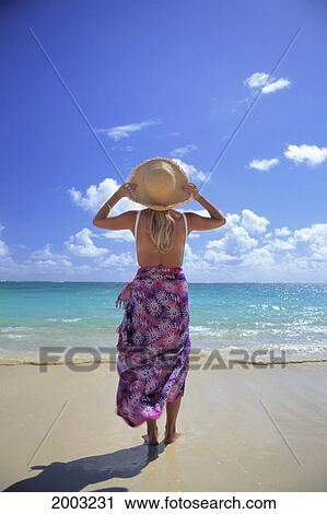 Stock Image - Back View Of Woman With Hat And Pareo At Beach Walks Towards Turquoise Ocean. Fotosearch