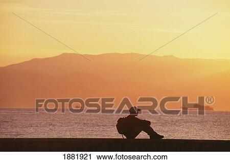 Stock Image - Backpacker In Silhouette Sitting By Coast. Fotosearch