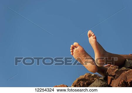 Picture - Bare feet hanging out over rocks against a blue sky; Oia, Santorini, Greece. Fotosearch