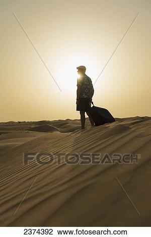 Stock Image - Barefoot man with suitcase on sand dune at dusk; Dubai, United Arab Emirates. Fotosearch