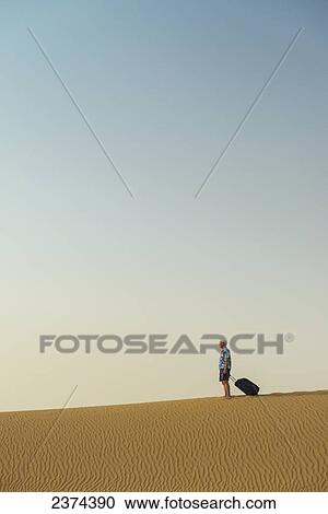 Stock Image - Barefoot man with suitcase on sand dune; Dubai, United Arab Emirates. Fotosearch