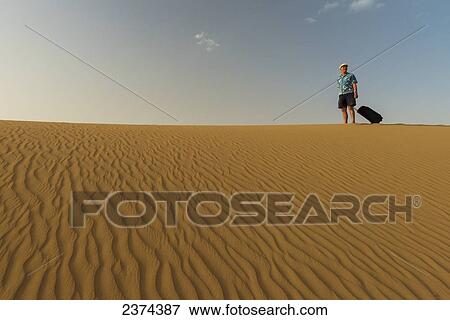 Stock Photo - Barefoot man with suitcase on sand dune; Dubai, United Arab Emirates. Fotosearch