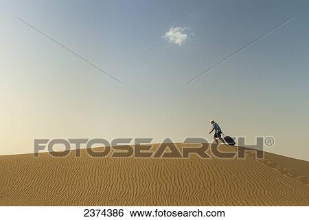 Stock Photograph - Barefoot man with suitcase walking on sand dune; Dubai, United Arab Emirates. Fotosearch