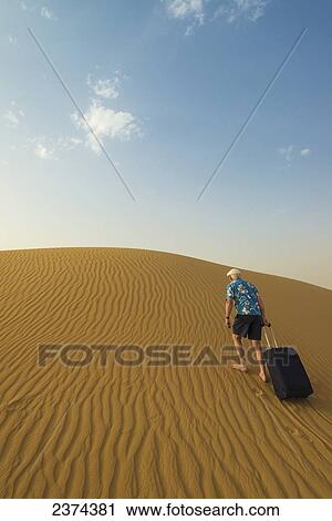 Stock Image - Barefoot man with suitcase walking up sand dune; Dubai, United Arab Emirates. Fotosearch