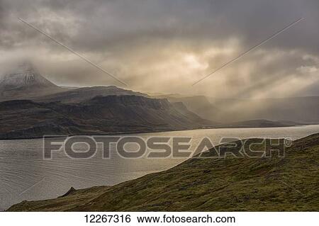 Stock Photograph - Beams of light shine down on the mountains and abandoned village of Djupavik; Iceland. Fotosearch