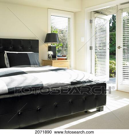 Stock Image - Bedroom With Black Leather Tufted Headboard And French Doors To Garden. Fotosearch