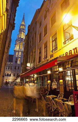 Stock Photography - Belgium, People walking down side street in front of restaurant with Town Hall behind in Grand Place at dusk; Brussels. Fotosearch
