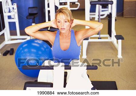 Picture - Blond Woman Sitting On Exercise Equipment Working On Her Abdominal Muscles. Fotosearch