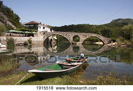 Stock Photo - Boats On River At Rijeka Crnojevica, Tributary Of Lake Skadar, With Old Stone Bridge In Distance. Fotosearch