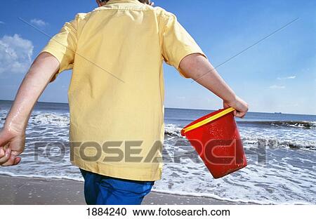 Stock Image - Boy Playing Along The Beach Carrying A Bucket. Fotosearch