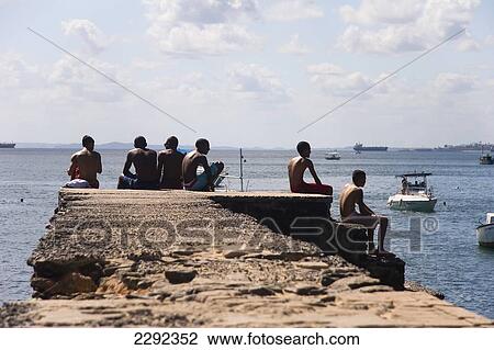 Stock Image - Brazil, Bahia State, Salvador, Porto da Barra Beach, Young men sitting on concrete pier. Fotosearch