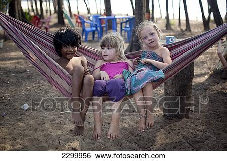 Stock Photograph - British Kids on holiday play in a hammock and make friends with a local girl on Turtle Beach, Goa, India.. Fotosearch