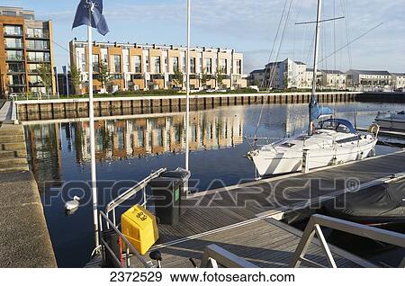 Stock Photo - Buildings reflected in the water along the shoreline and a sailboat mooring in the harbour; Portishead, North Somerset, England. Fotosearch