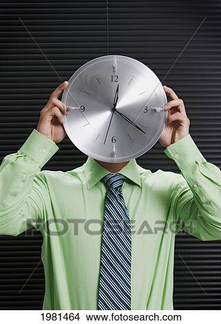Picture - Businessman In Office Holding A Silver Wall Clock In Front Of His Face.. Fotosearch