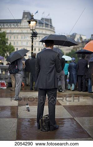 Stock Image - Businessman Standing In Rain With Umbrella In Trafalgar Square; London, England. Fotosearch