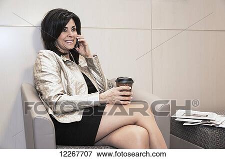 Stock Photo - Businesswoman talking on cell phone with coffee in office waiting area; Edmonton, Alberta, Canada. Fotosearch