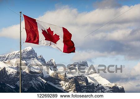 Stock Photo - Canadian flag blowing in the wind on a flag pole with snow covered mountain range in the background with blue sky and clouds; Canmore, Alberta, Canada. Fotosearch