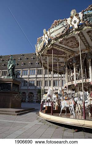 Stock Image - Carousel In The Town Square With A Bronze Statue And Blue Sky; Strasbourg, France. Fotosearch