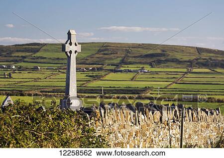 Stock Image - Celtic cross with stone fence and hilly grass fields in the distance with blue sky; Cross, County Clare, Ireland. Fotosearch