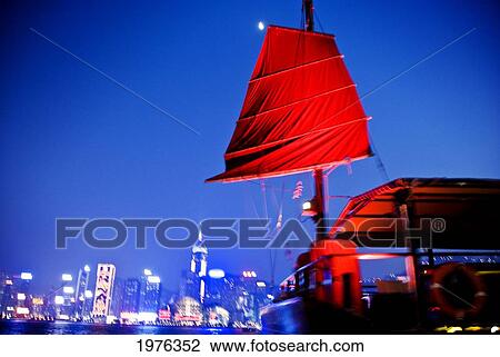 Stock Image - China, Tsim Sha Tsui K; Hong Kong, Red Sailed Chinese Junk Boat Passing In Front Of Hong Kong Island As Viewed From Promenade. Fotosearch
