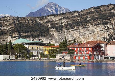 Stock Image - Cliffs and buildings along the shoreline of Lake Garda with men fishing in the harbour; Torbole, Trentino, Italy. Fotosearch