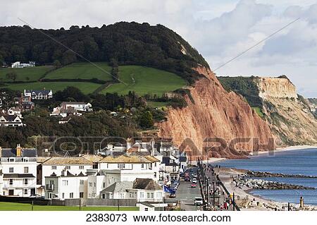 Stock Image - Cliffs and houses along the coast with pedestrians walking on a promenade along the water's edge; Sidmouth, Devon, England. Fotosearch