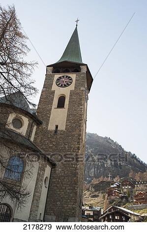 Stock Photo - Clock Tower And Cross Of A Church With Houses On The Hill In The Background; Zermatt Valais Switzerland. Fotosearch