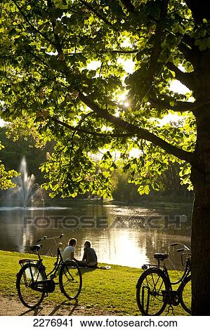 Stock Image - Couple relaxing beside lake in the Vondelpark in the evening; Amsterdam, Holland. Fotosearch