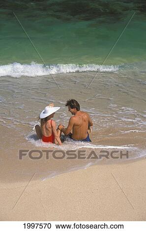 Stock Image - Couple Sit On Beach In Shallow Water White Hat Hawaii. Fotosearch