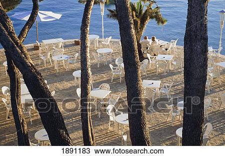 Stock Image - Couple Sitting At Outdoor Tables On Terrace Behind Trees At Sunset, Elevated View. Fotosearch