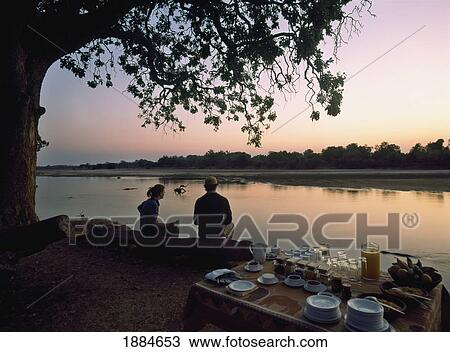 Stock Image - Couple Sitting On Log With Hot Drinks Before Dawn. Fotosearch