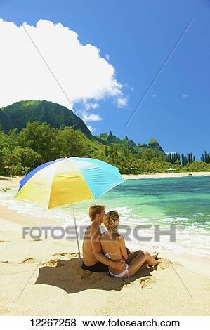Stock Photo - Couple sitting under an umbrella at the water's edge; Kauai, Hawaii, United States of America. Fotosearch