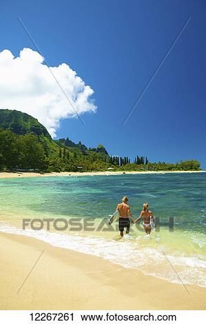 Stock Image - Couple walking into water with bodyboards; Kauai, Hawaii, United States of America. Fotosearch