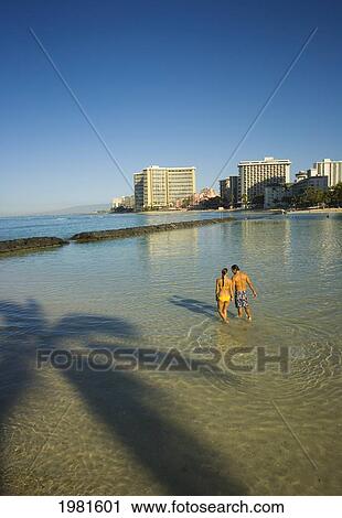 Stock Image - Couple walking through shallow water, Waikiki Beach; Waikiki, Oahu, Hawaii, United States of America. Fotosearch