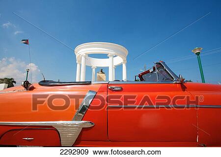 Stock Photo - Cuba, Side view of red vintage car with statue of Ernest Hemingway in background; Cojimar. Fotosearch