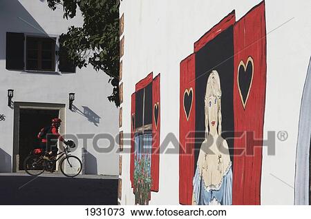 Stock Image - Cyclist at Kranjska Gora town near a painted building with fake windows and milk maid. Gorenjska Alpine region of Slovenia., Gorenjska Alpine region of Slovenia.. Fotosearch