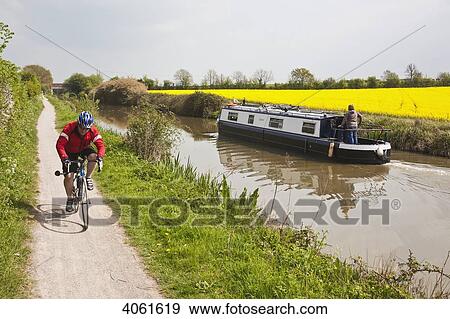 Cyclist cycling along canal side in open countryside passing narrowboat and Rape oilseed fields; Wiltshire, England View Large Photo Image Stock Photo - Cyclist cycling along canal side in open countryside passing narrowboat and Rape oilseed fields; Wiltshire, England. Fotosearch