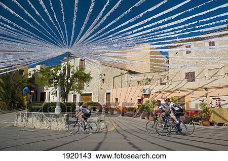 Stock Image - Cyclists Going Past Square With White Bunting In Caimari, Majorca, Spain. Fotosearch