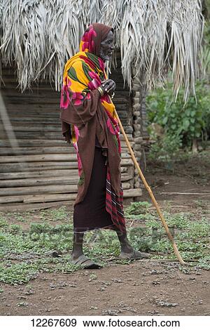 Stock Photo - Datoga elder in brightly coloured robe leans on cane in front of hut near Lake Eyasi; Tanzania. Fotosearch